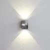 A square, metallic wall light fixture mounted on a textured white wall, casting light upwards and downwards. Voir les images du produit