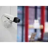 A white security camera is mounted on a white wall, with a blurred background of red structures and displays. Podgląd zdjęcia produktu