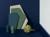 A still life arrangement of geometric shapes, two small vases, and an artichoke against a dark blue background. Produktbild-Vorschau