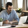 A man wearing a headset is working on a laptop at a desk. Produktbild-Vorschau