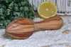 A wooden citrus reamer is placed on a sandy surface, next to a sliced lemon and a green plant. Produktbild-Vorschau