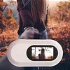 A person holding a small white camera with a screen displaying a woman's back, in an outdoor setting with trees and sunlight. Produktbild-Vorschau