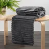 A folded gray blanket with a ribbed texture is draped over a light-colored wooden table. A potted plant with green leaves is visible on the left side of the table. Produktbild-Vorschau