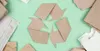 A recycling symbol made of folded brown paper is placed in the center of a light green background, surrounded by various pieces of crumpled and folded brown paper. Produktbild-Vorschau