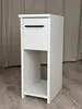 A white, narrow nightstand with a drawer and an open shelf, placed on a wooden floor in front of a beige curtain. Náhľad obrázku produktu