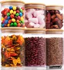 Sechs klare Glas-Lebensmittelaufbewahrungsbehälter mit Bambusdeckeln, gefüllt mit verschiedenen Trockenwaren wie Bonbons, Marshmallows, Datteln, Nudeln, Bohnen und Linsen, mit einem weißen Kreidestift im Vordergrund Produktbild-Vorschau