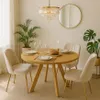 A dining room scene with a round wooden table set for a meal, surrounded by four upholstered chairs. A chandelier hangs above, and plants are placed in the background. Produktbild-Vorschau