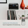 A retro-style record player with a CD player and radio is displayed on a shelf above a collection of vinyl records. A remote control is in the foreground. Produktbild-Vorschau