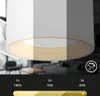 A close-up of a white cylinder-shaped lamp with a ribbed interior, showing different brightness levels. The background is blurred, showing a living room setting. Text and a slider bar are present at the bottom. Náhled obrázku produktu