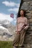 A woman leans against a stone wall in a mountainous landscape, with a flag in the background. Produktbild-Vorschau