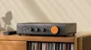 A dark gray Fosi Audio ZP3 HiFi preamplifier with an orange dial sits on a wooden cabinet. Produktbild-Vorschau