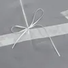 A close-up of a white ribbon tied in a bow on a grey fabric with a white stripe and a silver geometric pattern. Produktbild-Vorschau