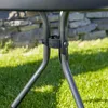 A close-up view of the legs and connecting mechanism of a dark-colored outdoor table, set on a green lawn with a stone wall in the background. Produktbild-Vorschau
