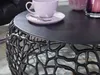 A close-up of a black coffee table with a decorative, organic base, featuring a purple cup and saucer with a spoon on top. Anteprima dell’immagine prodotto