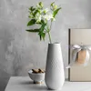 A white vase with white flowers sits on a marble table next to a gift box and a bowl of cookies. Produktbild-Vorschau