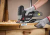 A person wearing protective gloves is using a cordless jigsaw to cut a piece of wood on a workbench. Produktbild-Vorschau