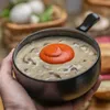 A hand holds a bowl of mushroom soup topped with a dollop of red sauce. In the background, there are mushrooms in a basket and some bread. Produktbild-Vorschau