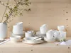 A collection of white porcelain dinnerware, including bowls, plates, and cups, arranged on a light wooden surface with a vase of white flowers in the background. Produktbild-Vorschau