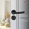 A close-up of a black door handle set on a white door, with a blurred background of a room interior. Produktbild-Vorschau