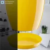 A yellow planter with a matching saucer is shown in a modern interior setting, with a kitchen island and stools visible in the background on the left, and a white geometric planter with a snake plant on the right. Podgląd zdjęcia produktu