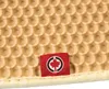 A close-up of a beige honeycomb-patterned mat with a small red tag featuring a black and white logo of a maple leaf inside a circle. Produktbild-Vorschau