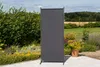 A dark gray privacy screen stands in a garden setting with plants and a paved area. Produktbild-Vorschau