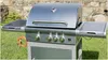 A stainless steel gas grill with side shelves and a gas hose attached, set up on a grassy lawn with a stone wall in the background. Podgląd zdjęcia produktu