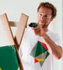 A man is using a cordless drill to assemble a wooden structure, possibly furniture. He is wearing safety glasses. Produktbild-Vorschau