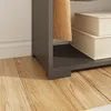 A close-up view of the bottom section of a gray wardrobe, showing its base and a shelf with two beige storage boxes on it. The floor is made of light brown wooden planks. Náhled obrázku produktu