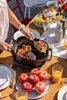 A table set for a meal outdoors, with a person serving a dish from an electric roaster. Anteprima dell’immagine prodotto