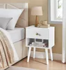 A white nightstand with a drawer and open shelf is placed next to a bed. A lamp and books are on top of the nightstand. Produktbild-Vorschau