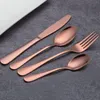 A set of copper-colored cutlery is arranged on a dark gray surface, with a white plate partially visible in the upper left corner. Voir les images du produit