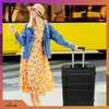 A woman stands next to a yellow bus, holding a suitcase. Anteprima dell’immagine prodotto