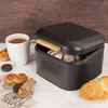 A black bread bin filled with bread, next to a cup of tea and some pastries on a wooden table. Produktbild-Vorschau
