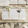 A white high-gloss coffee table with gold accents is placed in a living room setting, next to a white sofa and on a patterned rug. Produktbild-Vorschau
