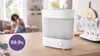 A baby sterilizer is shown on a kitchen counter, with a blurred background of a living room where a mother and child are visible. Produktbild-Vorschau