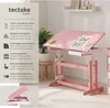 A pink and white children's desk with adjustable height and tiltable surface, shown in a room setting with a plant and a stool. Produktbild-Vorschau