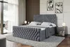 A bedroom scene featuring a dark gray upholstered box spring bed with a tufted headboard and footboard, set against a wall with wooden paneling and abstract art. A bedside table with lamps and books is also visible. Produktbild-Vorschau