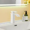 A white faucet is shown on a white sink, with a yellow wall and a mirror in the background. Náhľad obrázku produktu