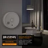 A smoke detector is shown on a gray wall, with a living room scene in the background at night. Produktbild-Vorschau