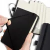A close-up shot of a black mini-notebook with its elastic band being stretched by a hand. Another notebook is visible in the background. Náhľad obrázku produktu