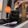 A person is pouring rice into a small black rice cooker on a stovetop, with a backdrop of water and sunset colors. Kitchen utensils are visible in the background. Produktbild-Vorschau