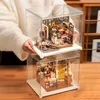 Two miniature dollhouse kits are stacked on top of each other, displayed in clear plastic cases on a wooden table. A person's hands are visible holding the top case. Podgląd zdjęcia produktu