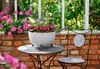 A white planter filled with pink and blue flowers sits on a small table next to a chair on a brick patio. Náhled obrázku produktu