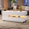 A modern living room with a white high-gloss coffee table in the foreground, featuring LED lighting and storage compartments. A brown sofa and a fireplace are visible in the background. Anteprima dell’immagine prodotto