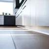 A low-angle shot of a modern kitchen with white cabinets and a light gray floor. Sunlight streams in from a window, casting striped shadows across the floor. Náhled obrázku produktu