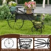 A black metal bicycle-shaped plant stand with three baskets holding potted plants, placed on a green lawn. Anteprima dell’immagine prodotto