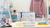 A sewing machine is displayed on a table with various sewing supplies and a pink shirt hanging in the background. Produktbild-Vorschau