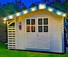 A white wooden shed decorated with string lights and a colorful fish ornament, set in a garden at dusk. Produktbild-Vorschau