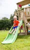 A young boy is sliding down a green slide attached to a wooden play structure in a grassy backyard. Produktbild-Vorschau
